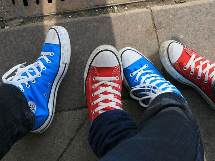 Two people standing on a sidewalk in high-top canvas sneakers, one pair bright blue and one pair red, both with white toe caps and laces