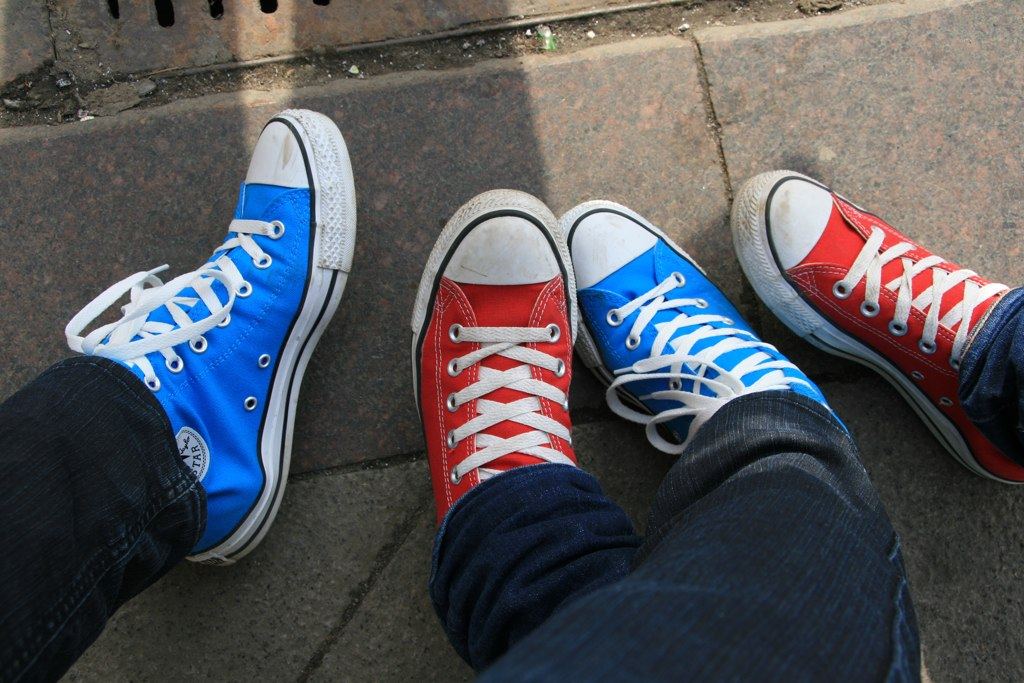 Two people standing on a sidewalk in high-top canvas sneakers, one pair bright blue and one pair red, both with white toe caps and laces
