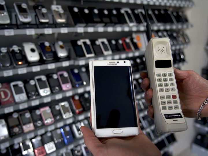 A hand holding a tall beige Motorola DynaTAC brick phone next to a modern touchscreen smartphone, with a wall of old mobile phones behind