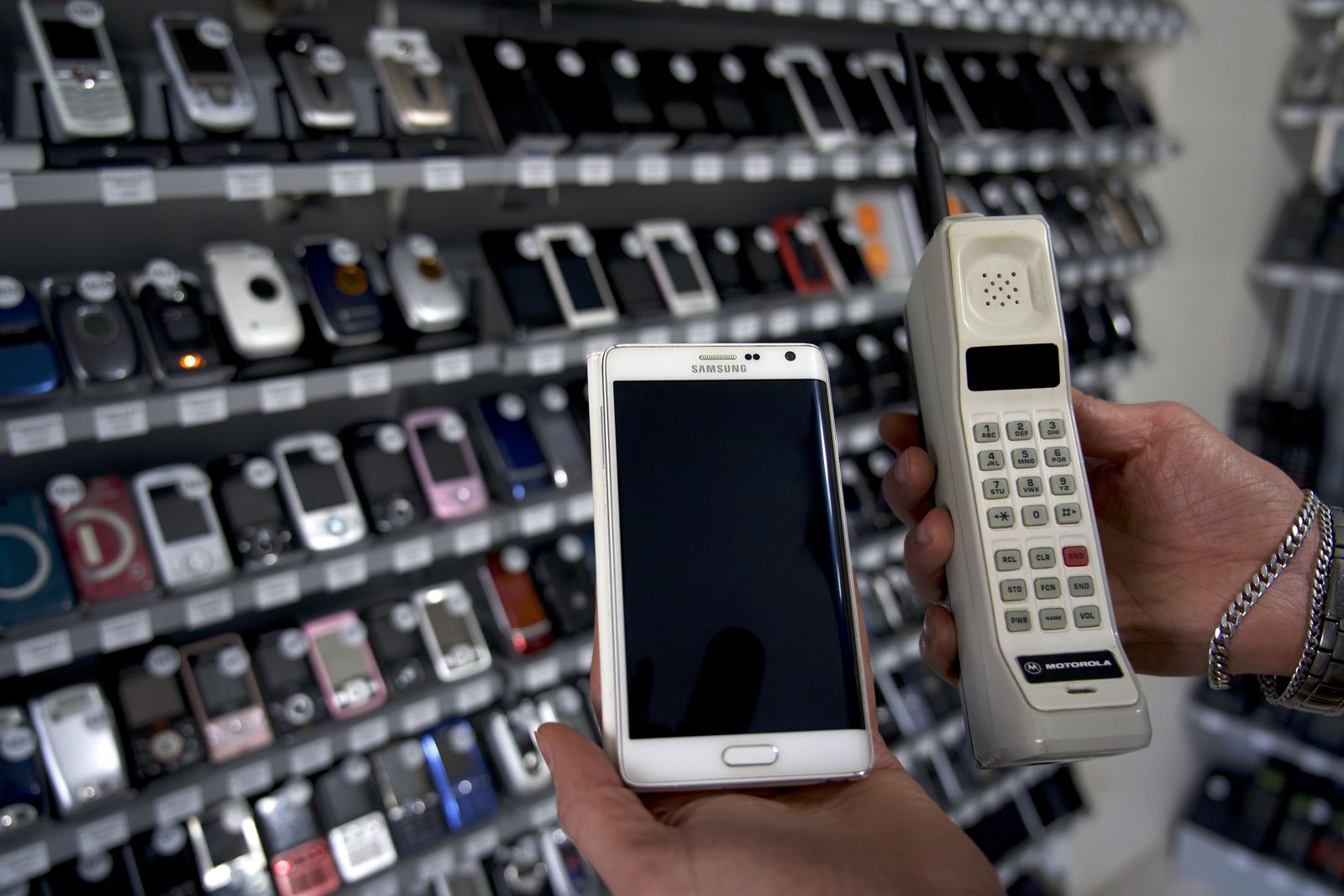 A hand holding a tall beige Motorola DynaTAC brick phone next to a modern touchscreen smartphone, with a wall of old mobile phones behind