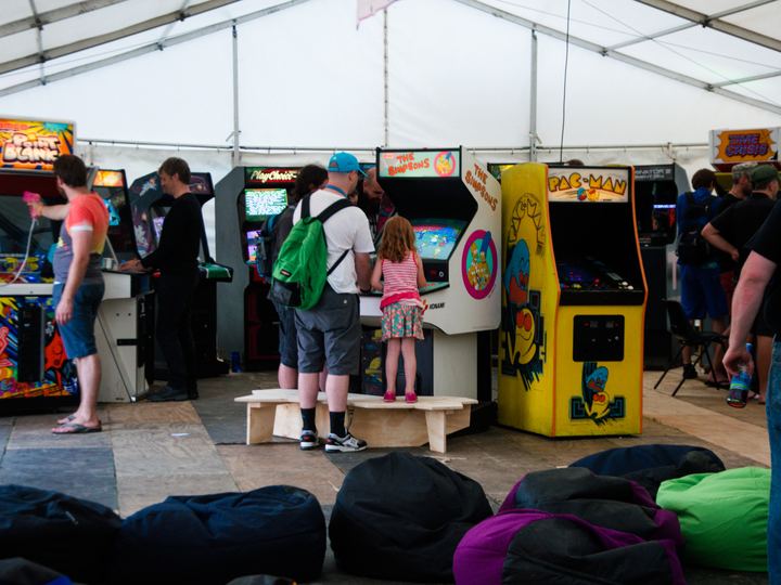 Visitors playing a row of classic upright arcade cabinets inside a tent at a retro gaming event, with beanbags in the foreground