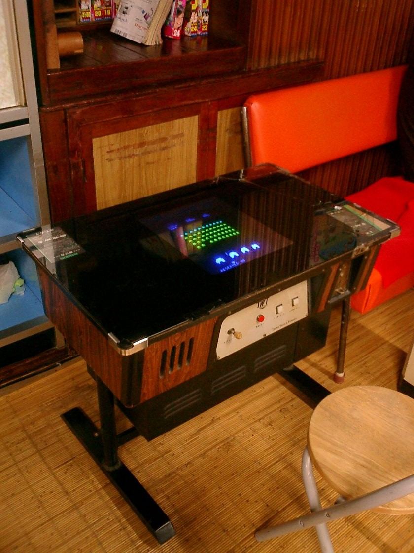 A Space Invaders cocktail table cabinet with a glass top over the glowing screen and a joystick panel