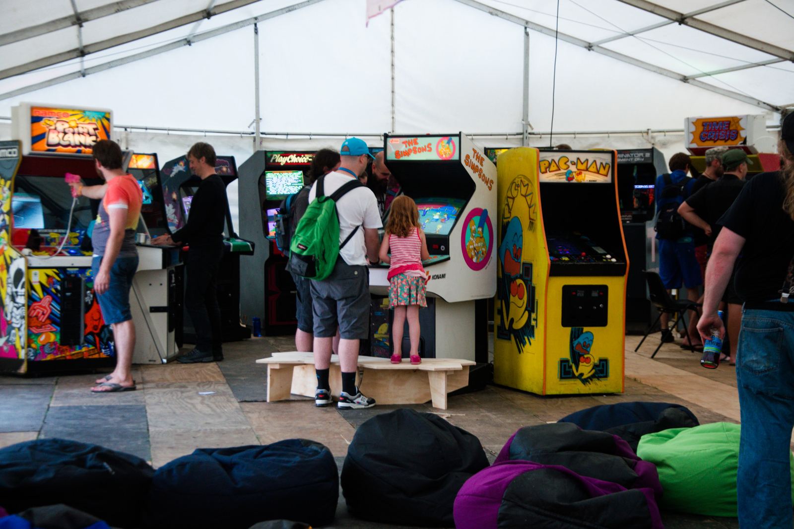 Visitors playing a row of classic upright arcade cabinets inside a tent at a retro gaming event, with beanbags in the foreground