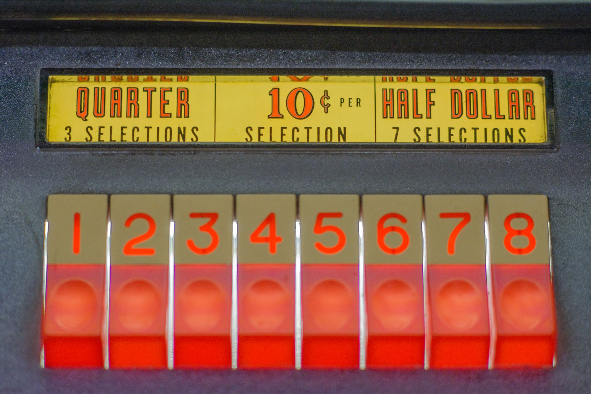 Close-up of glowing red numbered selection buttons on a vintage Seeburg jukebox, under a yellow price panel reading quarter, 10 cents per selection and half dollar