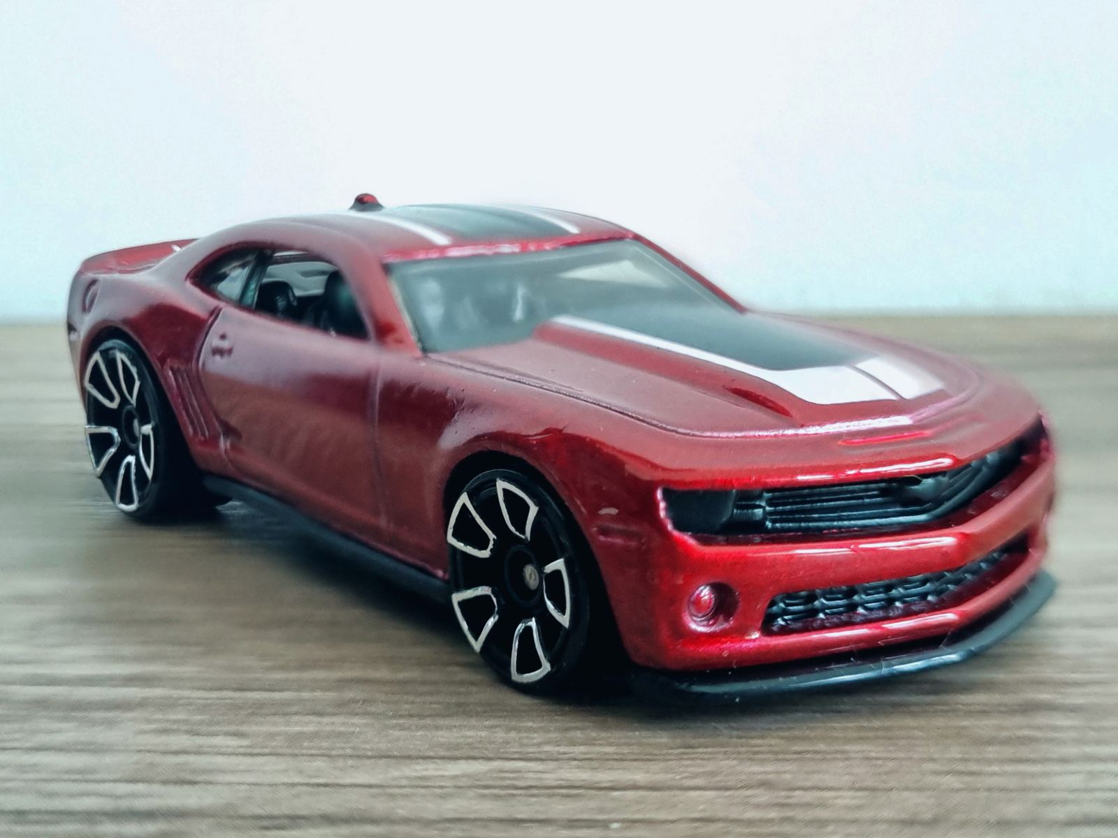 A small metallic red die-cast Chevrolet Camaro toy car with white racing stripes and black wheels on a wooden table