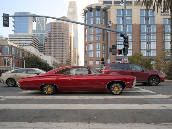 A deep red classic Chevrolet Impala lowrider with gold wire wheels sitting low at a crosswalk on the Embarcadero in San Francisco
