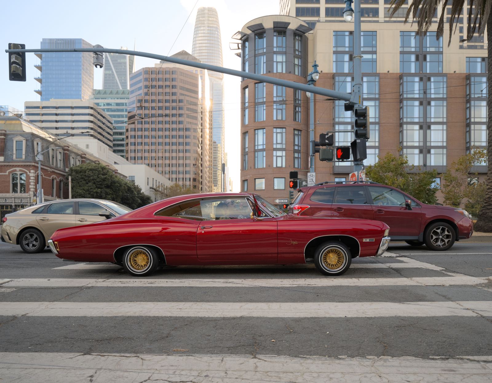 A deep red classic Chevrolet Impala lowrider with gold wire wheels sitting low at a crosswalk on the Embarcadero in San Francisco