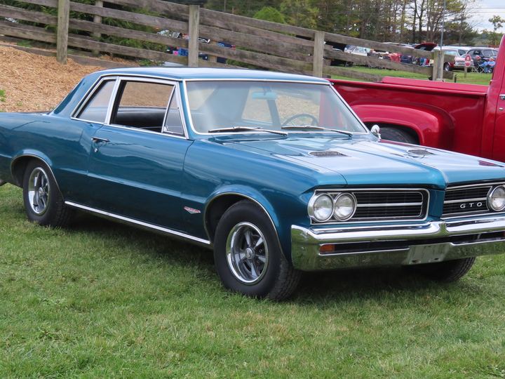 A teal blue 1964 Pontiac GTO coupe with chrome bumpers and a hood scoop parked on grass at a car show