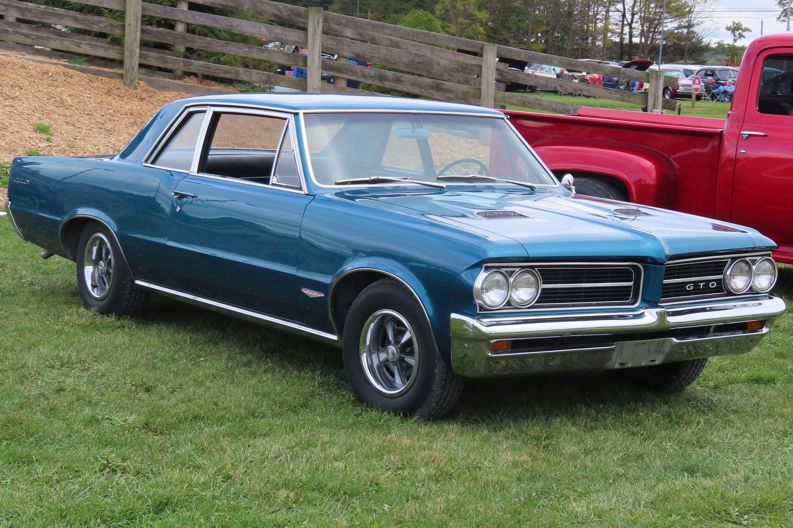 A teal blue 1964 Pontiac GTO coupe with chrome bumpers and a hood scoop parked on grass at a car show