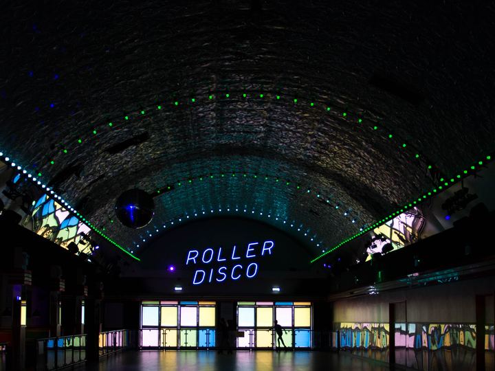 A dark arched roller rink hall lit with green laser lights, a mirror ball and a glowing neon Roller Disco sign