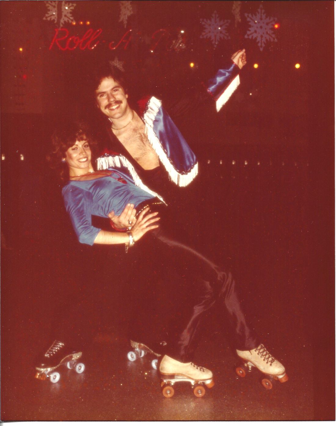 A 1970s skating couple in sparkling disco outfits posing mid-move on quad roller skates in a Brooklyn rink