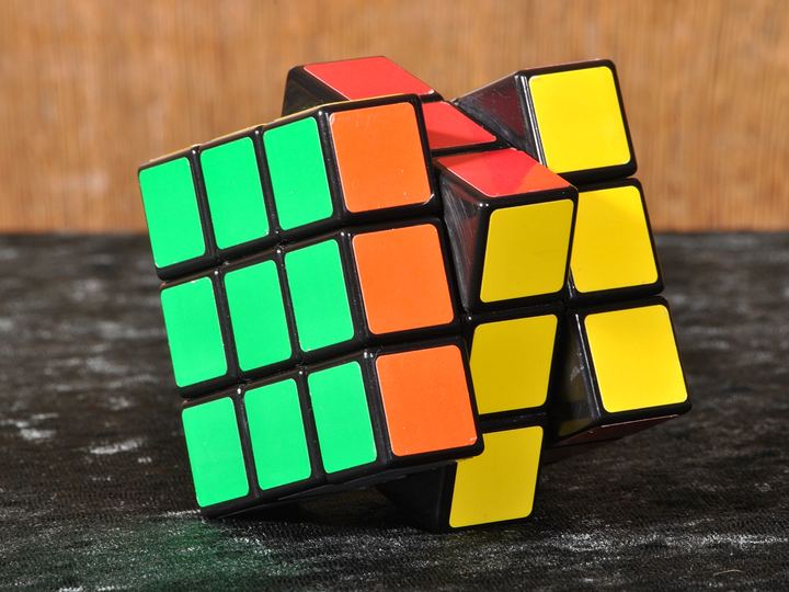 A classic Rubik's Cube mid-twist, with green, orange, yellow and red faces, sitting on a dark speckled surface