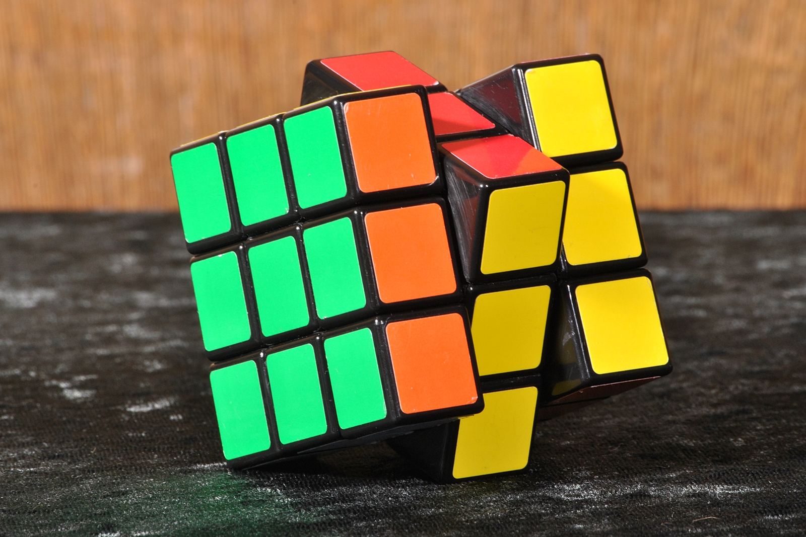 A classic Rubik's Cube mid-twist, with green, orange, yellow and red faces, sitting on a dark speckled surface